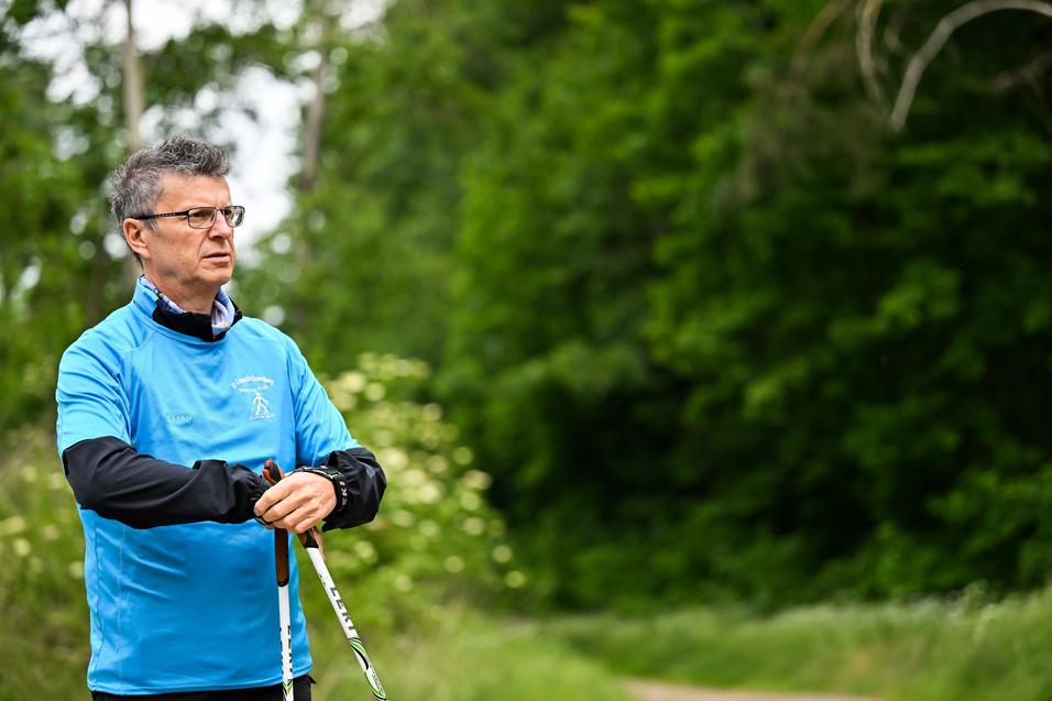 Mann in blauer Sportjacke hält Nordic Walking-Stöcke in einer grünen, bewaldeten Umgebung.