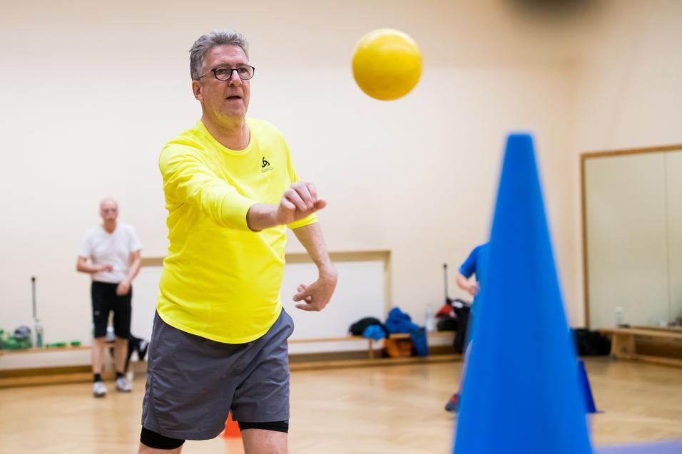 Ein Mann in gelber Sportkleidung wirft einen Ball auf einen blauen Kegel in einem Übungsraum. Hintergrund unscharf.