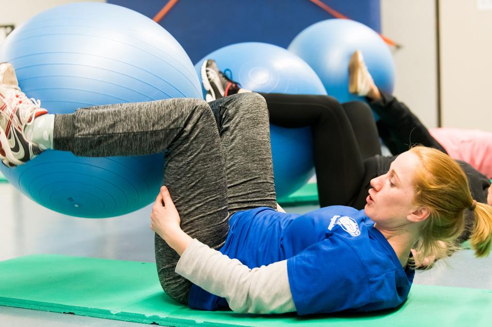 Frau trainiert auf einer Gymnastikmatte mit einem großen blauen Ball und hebt die Beine an, um die Muskulatur zu stärken.