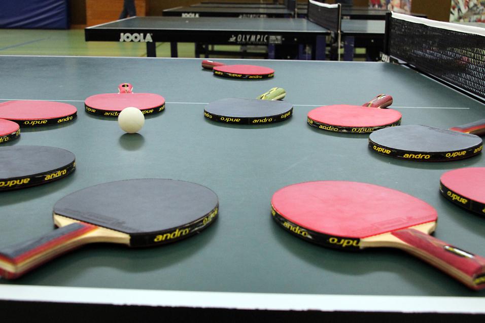 Mehrere Tischtennisschläger in verschiedenen Farben und ein Tischtennisball liegen auf einer grünen Tischtennisplatte.