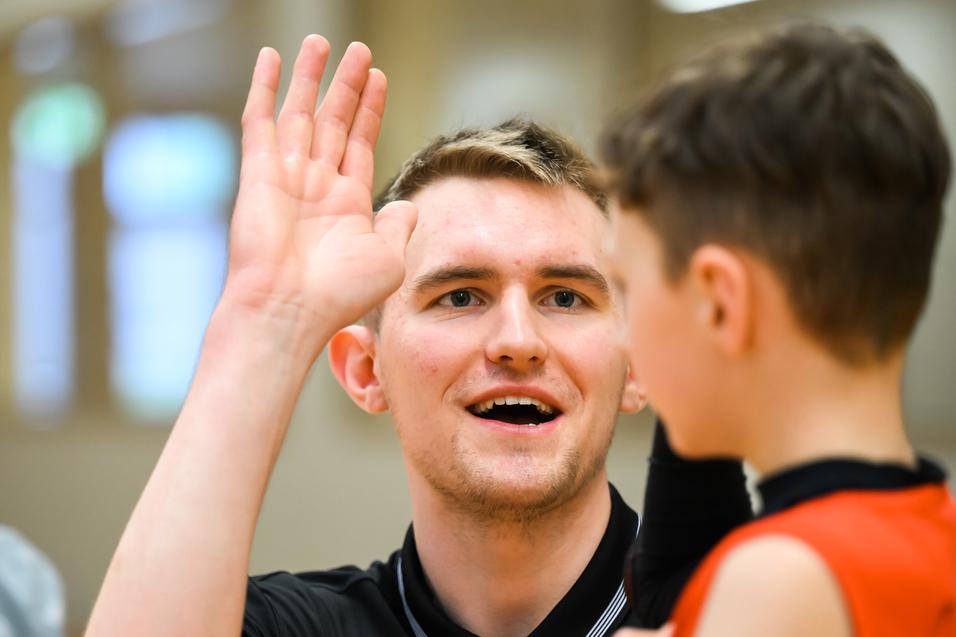 Trainer spricht mit einem jungen Spieler, während er seine Hand hebt, in einer Sporthalle mit Holzwand.