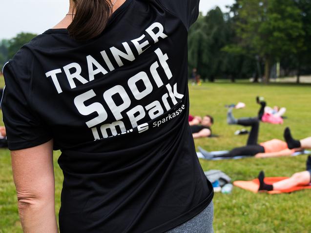 Trainerin mit schwarzem Shirt leitet Sportübungen im Park; Teilnehmer liegen auf Matten und trainieren im Hintergrund.