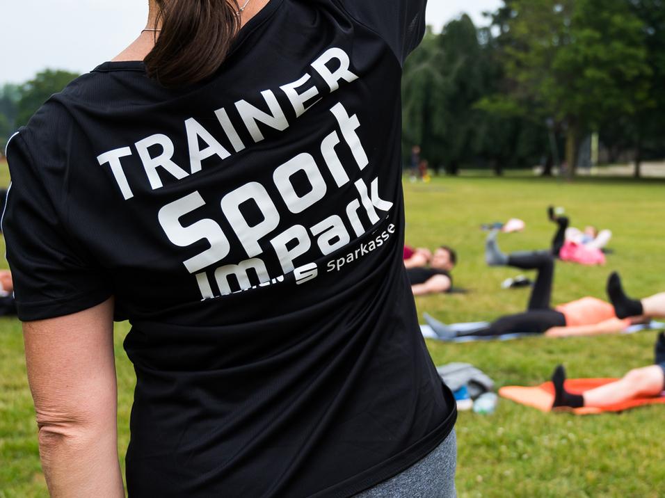 Trainerin mit schwarzem Shirt leitet Sportübungen im Park; Teilnehmer liegen auf Matten und trainieren im Hintergrund.